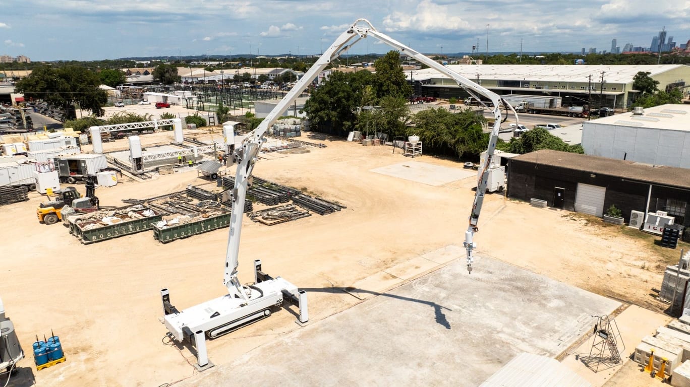 Aerial view of an ICON Titan concrete printing system on a job site, its robotic arm extended over a concrete slab with a city skyline behind.