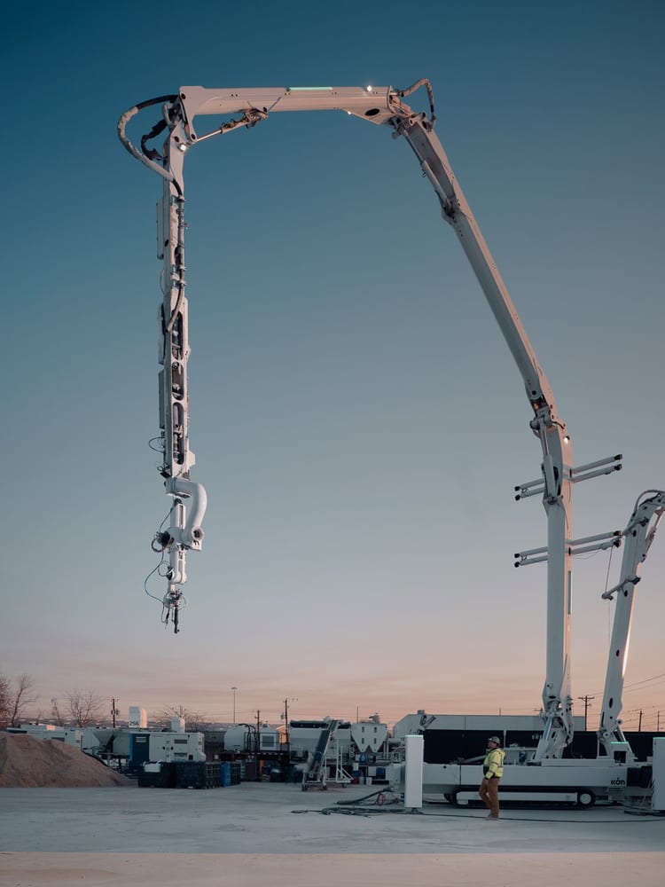ICON's Titan Print System at dusk, its robotic arm extended upward beside a worker and support equipment.