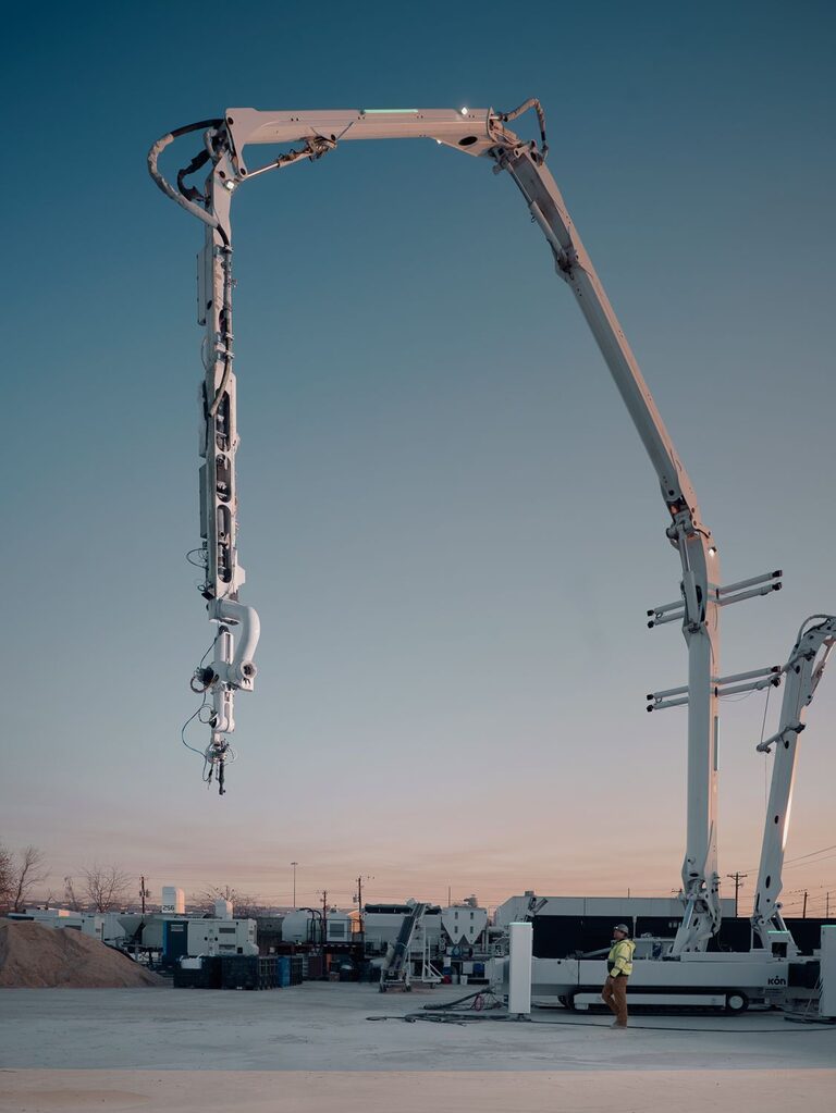 ICON's Titan Print System at dusk, robotic arm extended beside a worker and support equipment.